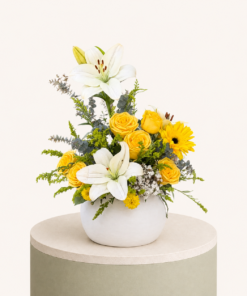 Luxury flower arrangement featuring yellow roses, white lilies, gerbera flowers, and fresh greenery in a white ceramic vase.