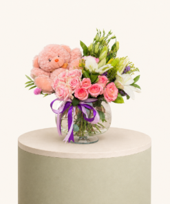 Elegant flower arrangement with pink roses, white lilies, and a soft pink teddy bear in a glass vase by Marino Flowers.