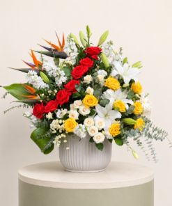 Luxury mixed flower arrangement with red roses, yellow roses, white lilies, birds of paradise, and eucalyptus in a ceramic vase.