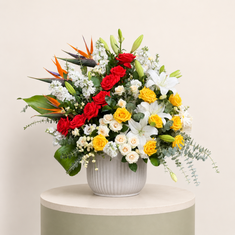Luxury mixed flower arrangement with red roses, yellow roses, white lilies, birds of paradise, and eucalyptus in a ceramic vase.