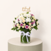 Get Well Soon flower arrangement with white chrysanthemums, cream spray roses, pink flowers, and الحمد لله على السلامة topper in a glass vase.