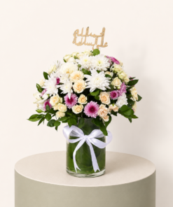 Get Well Soon flower arrangement with white chrysanthemums, cream spray roses, pink flowers, and الحمد لله على السلامة topper in a glass vase.