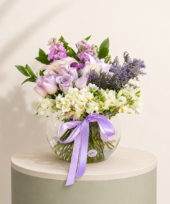 Lavender rose flower arrangement with white freesia, lilies, stock flowers, and greenery in a glass vase.