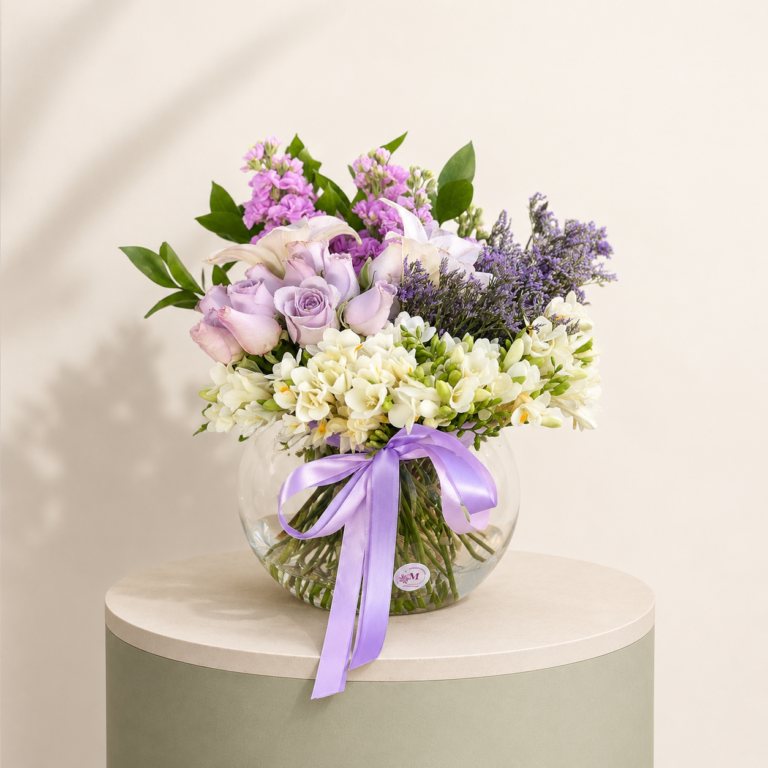 Lavender rose flower arrangement with white freesia, lilies, stock flowers, and greenery in a glass vase.