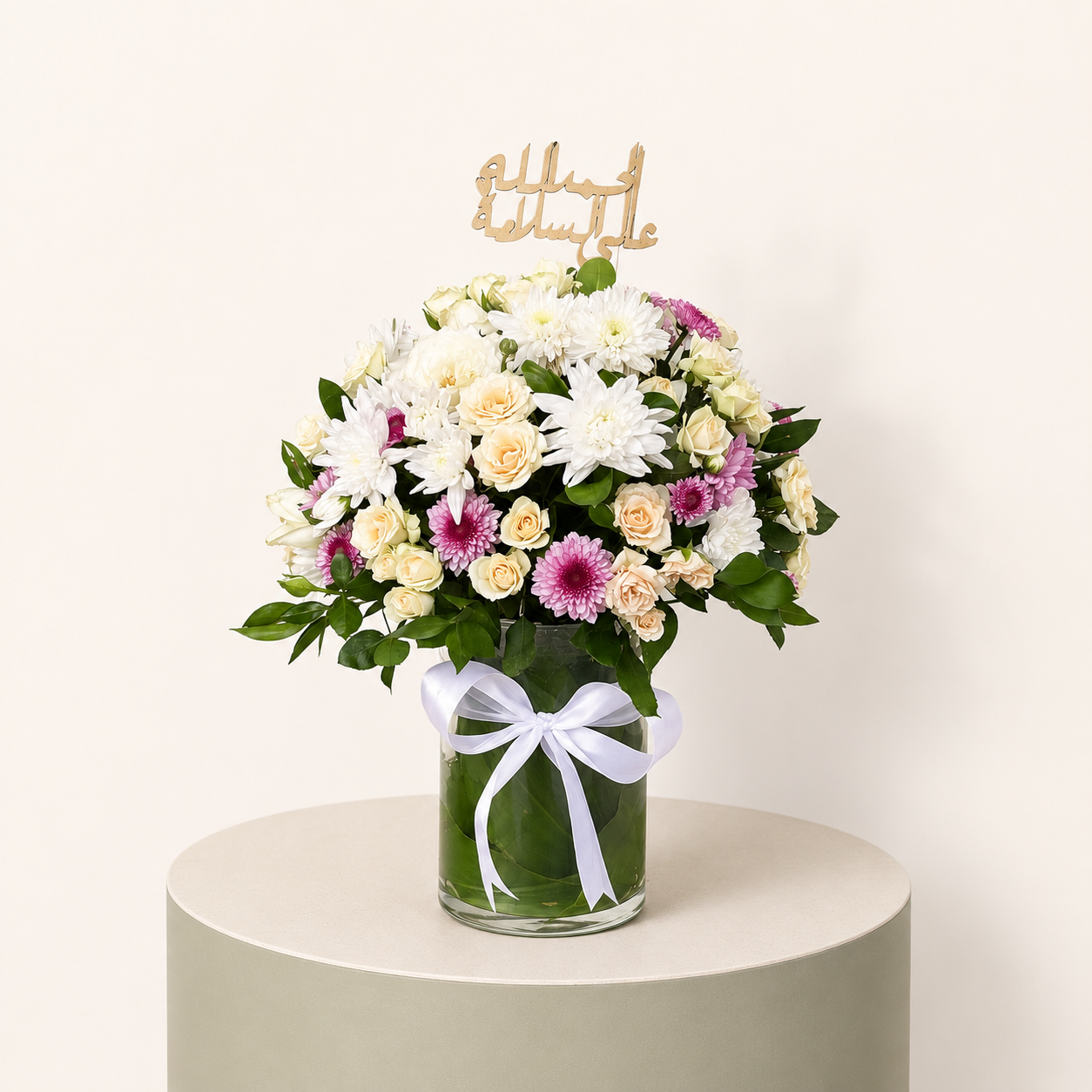 Get Well Soon flower arrangement with white chrysanthemums, cream spray roses, pink flowers, and الحمد لله على السلامة topper in a glass vase.