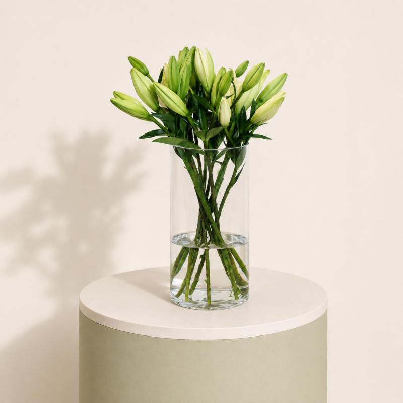 White lily stems arranged in a clear glass vase on a soft beige pedestal by Marino Flowers