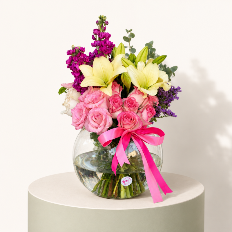 Pink roses, white lilies, purple stock flowers, and greenery arranged in a clear glass bowl by Marino Flowers.