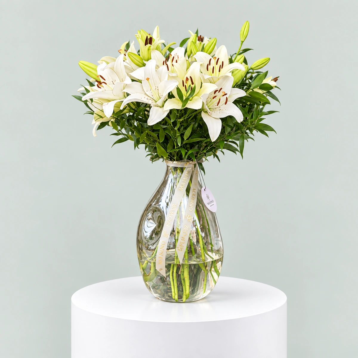 Pure Majesty – 10 White Lily Stems in Luxury Glass Vase Luxury arrangement of 10 white lily stems with open blooms and green buds in a large clear glass vase.