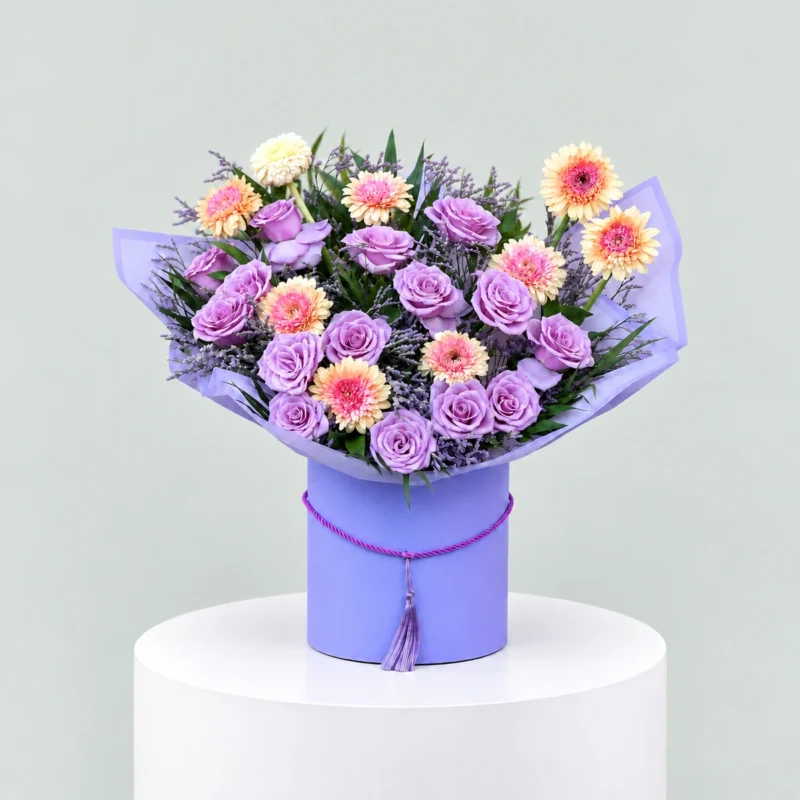 Lavender roses and peach-pink gerbera arranged with purple filler flowers in a lavender cylindrical flower box.