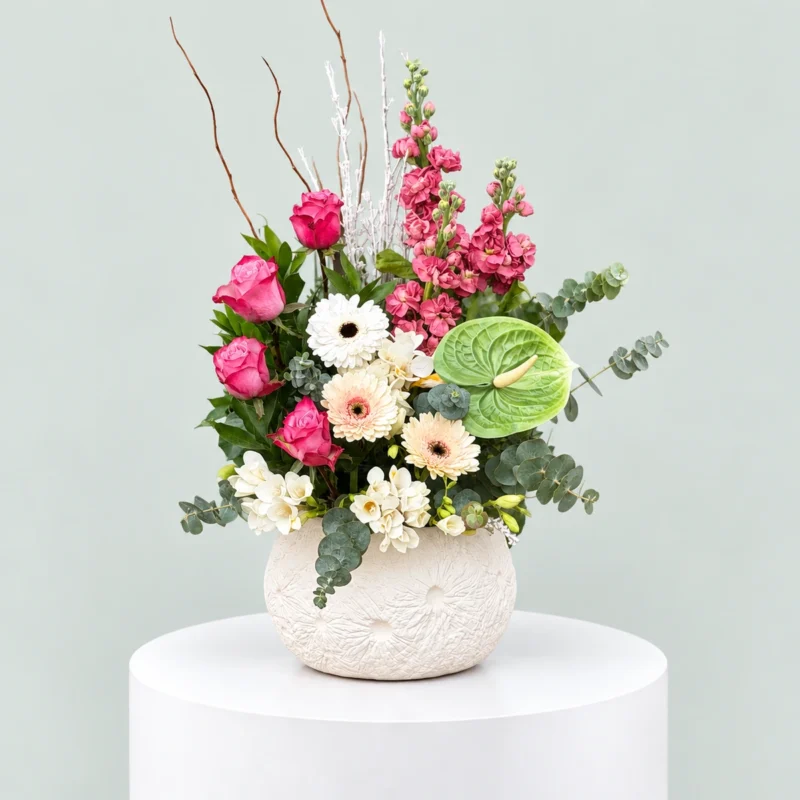 Pink roses, pink stock flowers, white gerberas and green anthurium with eucalyptus in a textured white ceramic vase.