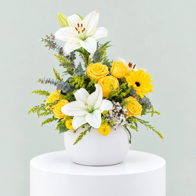 White lilies, yellow roses and yellow gerbera daisies arranged with eucalyptus and greenery in a round white ceramic vase.